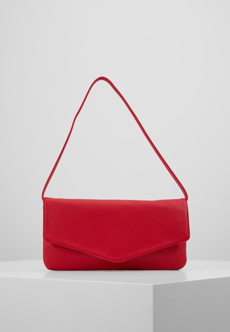 Červená texturovaná kožená kabelka ve stylu obálky s tenkým popruhem vystavená na bílém podstavci na šedém pozadí.
