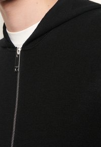 Svart zip-up hoodie med fin stickad textur, metallblixtlås och huva. Rundad halsringning över ett vitt baslager.