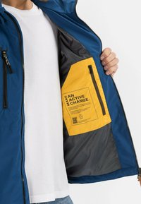 Blaue Jacke mit einer gelben Innentasche. Die Tasche hat Text und einen QR-Code. Glatte Textur, mit Reißverschlüssen und praktischer Gestaltung.