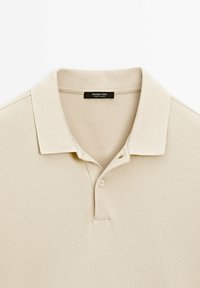 Polo beige avec un col côtelé et une patte de boutonnage à deux boutons, avec une étiquette noire indiquant « Massimo Dutti Made in Turkey ».