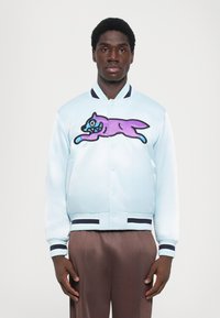  Lichtblauwe satijnen bommervest met een grote paarse en teal grafiek van een springende kat. Ribgebreide marineblauwe manchetten en zoom.