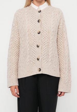 Femme portant un cardigan beige en maille torsadée boutonné, par-dessus une chemise blanche à col et un pantalon noir, debout devant un fond uni.