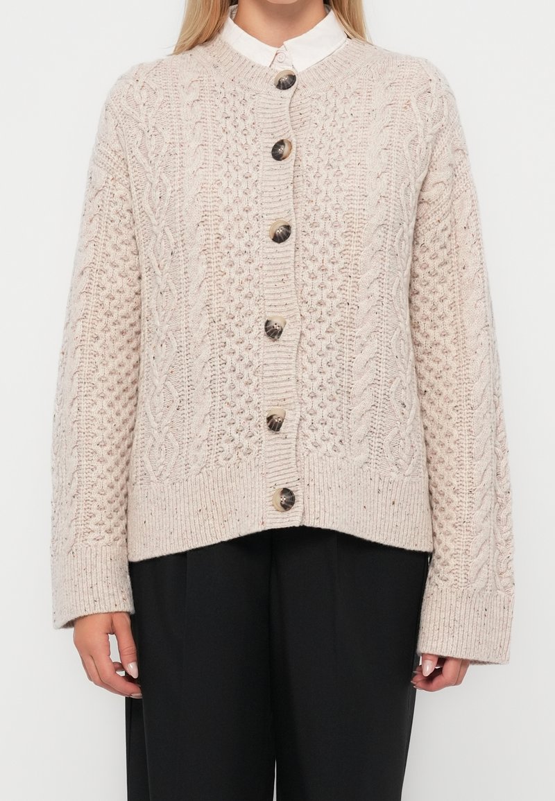 Femme portant un cardigan beige en maille torsadée boutonné, par-dessus une chemise blanche à col et un pantalon noir, debout devant un fond uni.