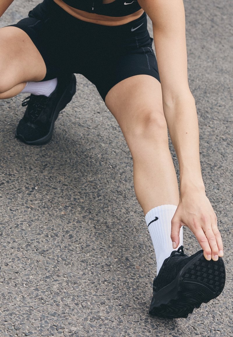 Personne portant des vêtements de sport noirs et des chaussettes blanches Nike étirant la jambe et touchant une chaussure de course noire sur une surface en asphalte.