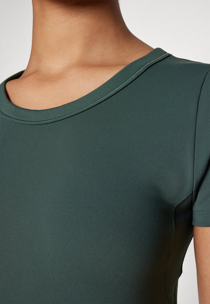 Grünes Sportoberteil mit rundem Ausschnitt, kurzen Ärmeln, glatter Textur und dezenten Nähdetails, vor einem schlichten Hintergrund präsentiert.