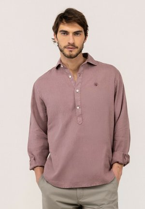 Hombre con cabello castaño y barba que lleva una camisa de manga larga color rosa polvo con tres botones, con las manos en los bolsillos de pantalones beige.