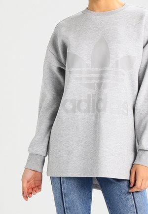 Persoon die een lichtgrijze Adidas-sweatshirt met een groot frontlogo en blauwe spijkerbroek draagt, staand tegen een effen witte achtergrond.
