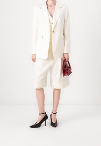 Ušitý bílý blejzr s odpovídajícími kraťasy, hladká textura. Černé lodičky s špičatou špičkou a členkovým páskem. Červená kabelka se strukturovaným designem.