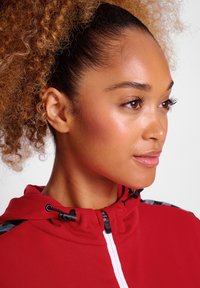 Rote Zip-Hoodie aus strukturiertem Stoff, schwarze Akzente an den Schultern und verstellbarer Halskordel. Nahaufnahme des Profils eines Modells.