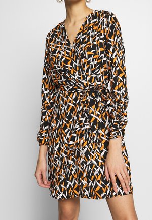 Nainen pitkähihainen kietaisumekko, jossa mustavalkoinen ja oranssi abstrakti lehtikuvio sekä solmittu vyötärönauha.