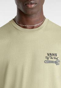 Gros plan d'une personne portant un t-shirt Vans vert clair avec un motif clé à molette et le texte « Off The Wall » sur la poitrine, ainsi qu'un collier chaîne en argent.