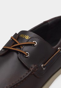 Καφέ σκούρο δερμάτινο καθημερινό παπούτσι με ταμπά κορδόνια περασμένα από μεταλλικά ρέλια και χρυσή επωνυμία "HUGO" στην περιοχή της γλώσσας.