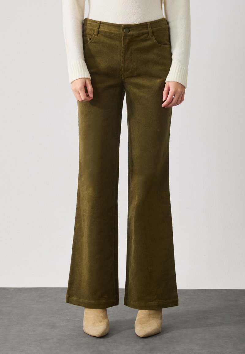 Femme portant un pantalon large en velours côtelé vert olive, un pull crème et des bottes à bouts pointus beige, debout sur un sol gris.