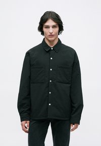 Camisa preta, abotoada com colarinho, duas bolsos frontais e um tecido texturizado. O design apresenta botões de pressão e um corte descontraído.