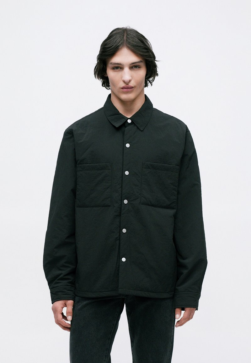 Camisa preta, abotoada com colarinho, duas bolsos frontais e um tecido texturizado. O design apresenta botões de pressão e um corte descontraído.
