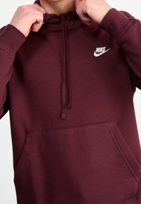 Bordó mikina Nike s kapucí na stažení, klokaní kapsou a bílým logem na hrudi. Vyrobená z měkké, texturované látky.