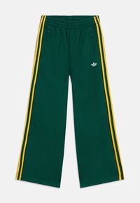 Grønne atletiske bukser i mesh med løs passform, med to glidelåslommer på sidene og gule striper langs sidene. Hvitt merke på forsiden.