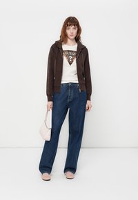 Bruine zip-up hoodie over een wit shirt met een driehoekig logo, gecombineerd met losse blauwe denim jeans en een lichtroze handtas.