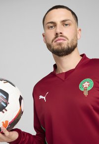 Rotes Sporttrikot mit grünen Akzenten und einem Wappen, kombiniert mit einem gemusterten Fußball. Der Stoff hat eine glatte Textur; das Logo ist hervorstechend platziert.