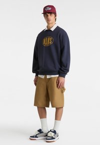 Sudadera de marina con logo amarillo, pantalones cortos beige y zapatillas azul/blancas. Gorro rojo y calcetines blancos añaden contraste al conjunto.