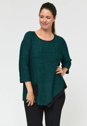 Teal getextureerd top met een ronde hals, driekwart mouwen en een zwarte zakdetails aan de linkerkant. In combinatie met zwarte broek.