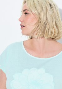 Maglietta corta a maniche corte verde menta chiaro con un grande disegno di fiore tratteggiato in bianco. Il collo presenta un bordo liscio a contrasto.