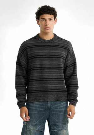 Giovane uomo con capelli ricci che indossa un maglione a maglia grigio scuro a righe e jeans cargo blu, in piedi davanti a uno sfondo bianco.