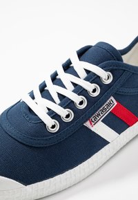 Blå lærreds sneakers med hvide snørebånd og gummisåle. Har røde og hvide stribede detaljer samt et mærke på siden. Tekstureret overflade.
