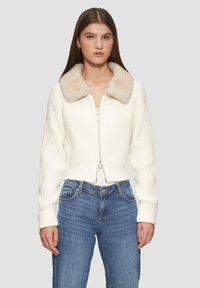 Gekürzter weißer Cardigan mit geripptem Muster, ausgestattet mit einem weichen, beigen Kunstfellkragen und einem zwei-Wege-Reißverschluss. Getragen mit blauen Jeans.