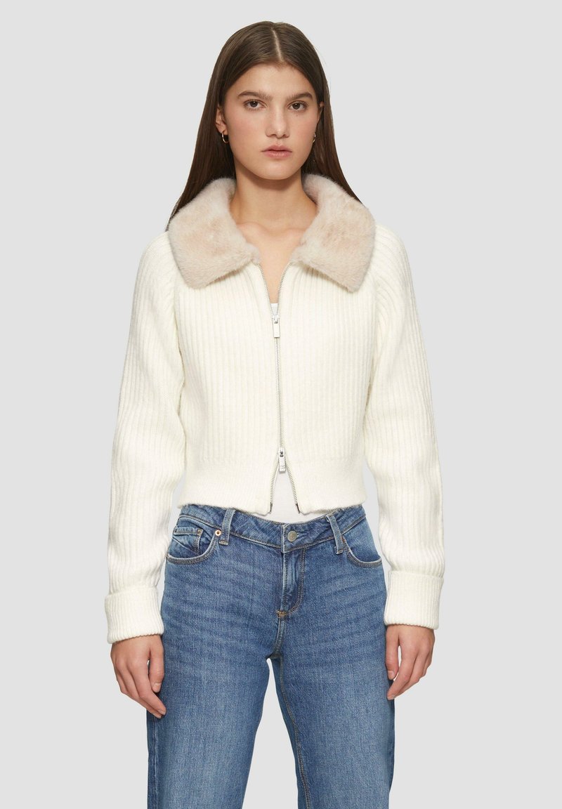 Gekürzter weißer Cardigan mit geripptem Muster, ausgestattet mit einem weichen, beigen Kunstfellkragen und einem zwei-Wege-Reißverschluss. Getragen mit blauen Jeans.