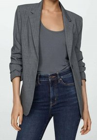 Blazer à fines rayures grises en tissu texturé, associé à un débardeur gris et un jean denim bleu foncé à coupe standard avec fermeture à boutons.