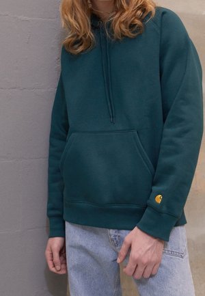 Person iført en mørk teal hættetrøje med frontlomme og lille gul logo på ærmet, kombineret med lyseblå jeans, stående op ad en grå væg.