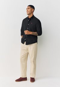 Črna srajca z gumbi z majhnim rdečim emblemom, bež hlače in škornji iz bordo semiša. Gladek material in sproščen kroj. Priložnostni slog.