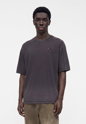 Jeune homme aux cheveux courts et bouclés portant un t-shirt uni sombre oversize et un pantalon marron clair, debout devant un fond blanc.