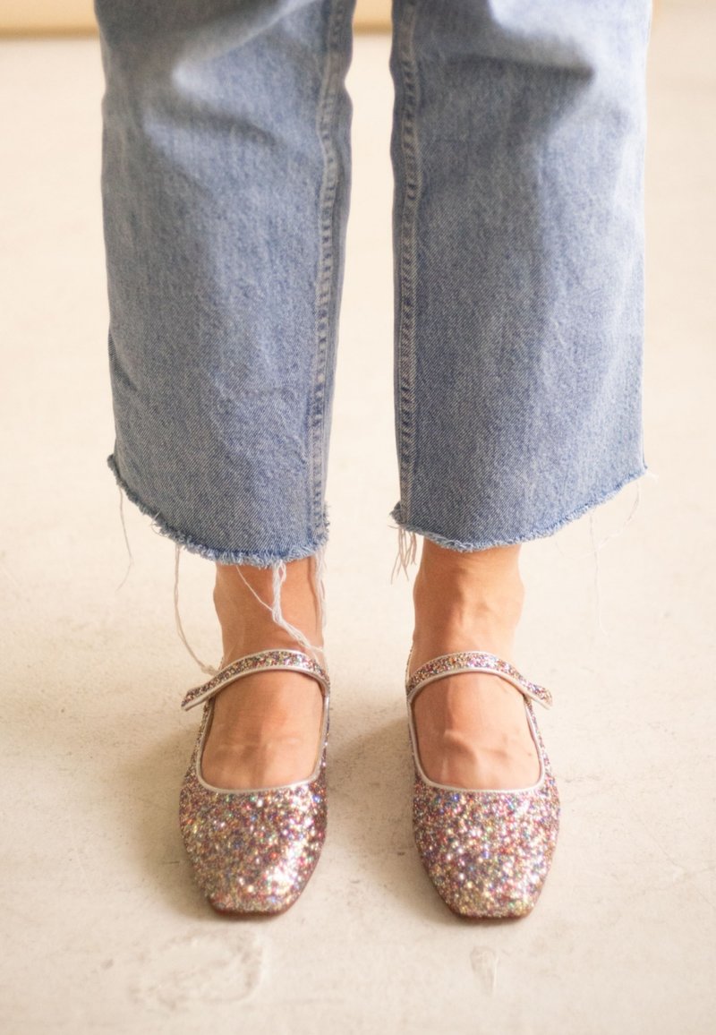 Pieds chaussés de chaussures Mary Jane pailletées multicolores étincelantes assorties à un jean bleu clair à ourlet effiloché sur un sol neutre.