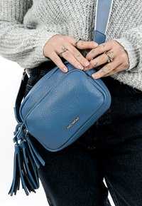 Maža mėlyna odinė crossbody rankinė su tekstūruotu paviršiumi, turinti užtrauktuką ir ilgas pūkuotas detales, dėvima prie pilko megztinio.