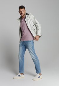 Lichtgrijze waterdichte jas met capuchon, gedragen over een roze poloshirt. Blauwe jeans en witte sneakers met marineblauwe accenten maken de outfit af.