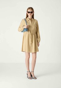 Femme portant une robe beige à boutons avec une ceinture à la taille, des lunettes de soleil noires, un sac à main bleu et des sandales à talons bleues, se tenant devant un fond uni.
