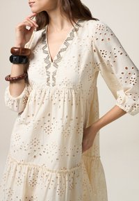 Vestido de crema claro con delicados recortes de ojal en forma de flor, escote en V profundo con costuras decorativas y dobladillo gather, combinado con pulseras de madera.