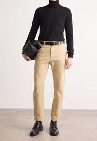 Personne portant un col roulé noir, un pantalon beige avec une ceinture noire, des chaussures noires, et tenant une pochette en cuir noir devant un fond uni.