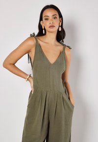 Olive green jumpsuit with adjustable tie straps, V-neck, pleated design, side pockets, and textured fabric.