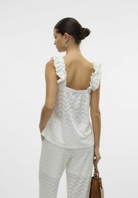 Witte top met ruches op de schouders, met een uitgesneden ontwerp en zigzagpatroon. Gecombineerd met bijpassende witte zigzagbroek en een bruine handtas.