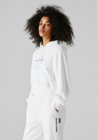 Vrouw met krullend haar, gekleed in een volledig witte hoodie en joggingbroek met de tekst "Red Bull Racing" op de broekzak, staand voor een grijze achtergrond.