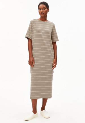 Femme portant une robe midi beige avec de fines rayures horizontales blanches et des chaussures blanches faciles à enfiler, debout devant un fond blanc uni.