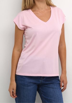 Kobieta w jasnoróżowej koszulce z krótkim rękawem i dekoltem w serek oraz niebieskich dżinsach, stojąca na tle jednolitego białego tła.