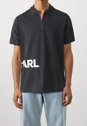 Mann trägt schwarzes kurzärmeliges Poloshirt mit Reißverschluss und weißem teilweisem "KARL"-Schriftzug nahe am Saum, kombiniert mit hellblauen Jeans.