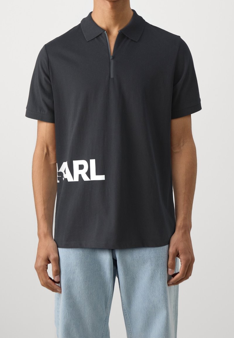 Homme portant un polo noir à manches courtes avec zip et texte partiel blanc "KARL" près de l'ourlet, associé à un jean en denim bleu clair.