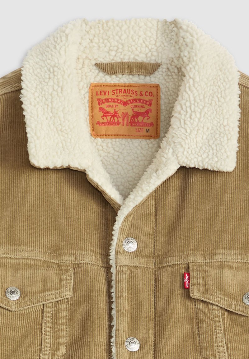 Chaqueta de pana en marrón con un cuello de borreguillo blanco. Presenta botones a presión de metal y una etiqueta roja de Levi's en la parte frontal. Talla M.