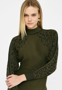 Olivno zelena pletena majica s visokim ovratnikom, ki ima teksturirano črno leopardo žgečkanje na rokavih in diskretne rebraste detajle.