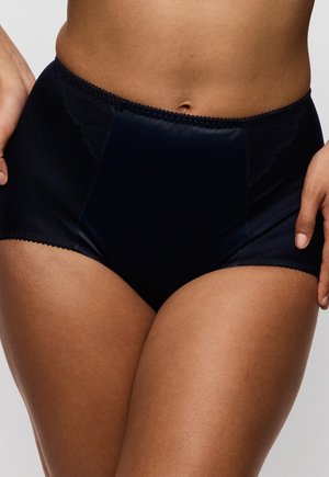 Vrouw gekleed in zwart satijnen lingerie met hoge taille en kanten details aan de zijkanten, staand met handen rustend nabij haar heupen.